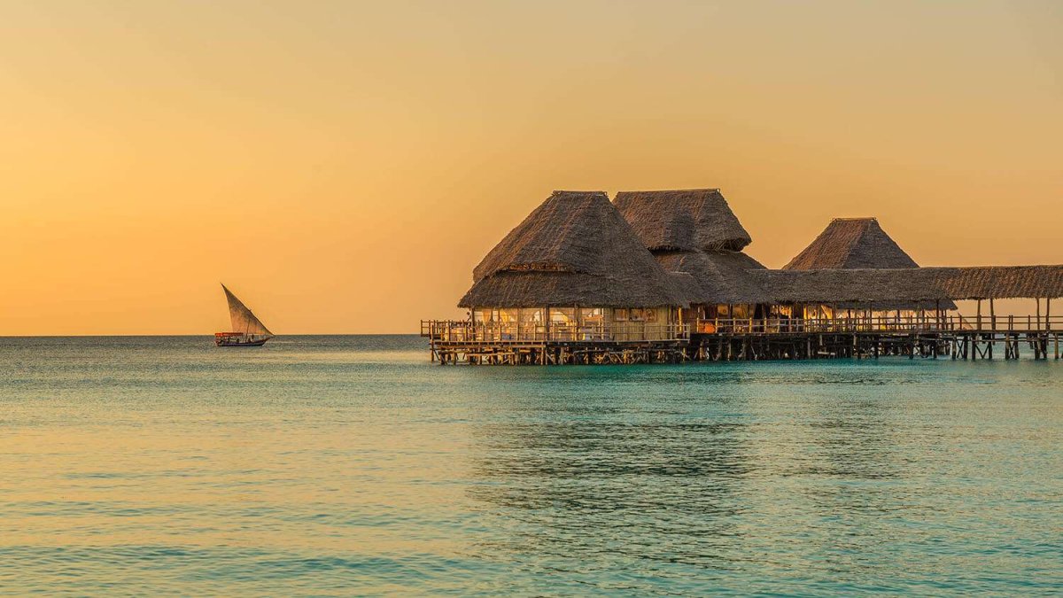 A thatched restaurant on stilts over the sea at sunset, with a dhow sailing past